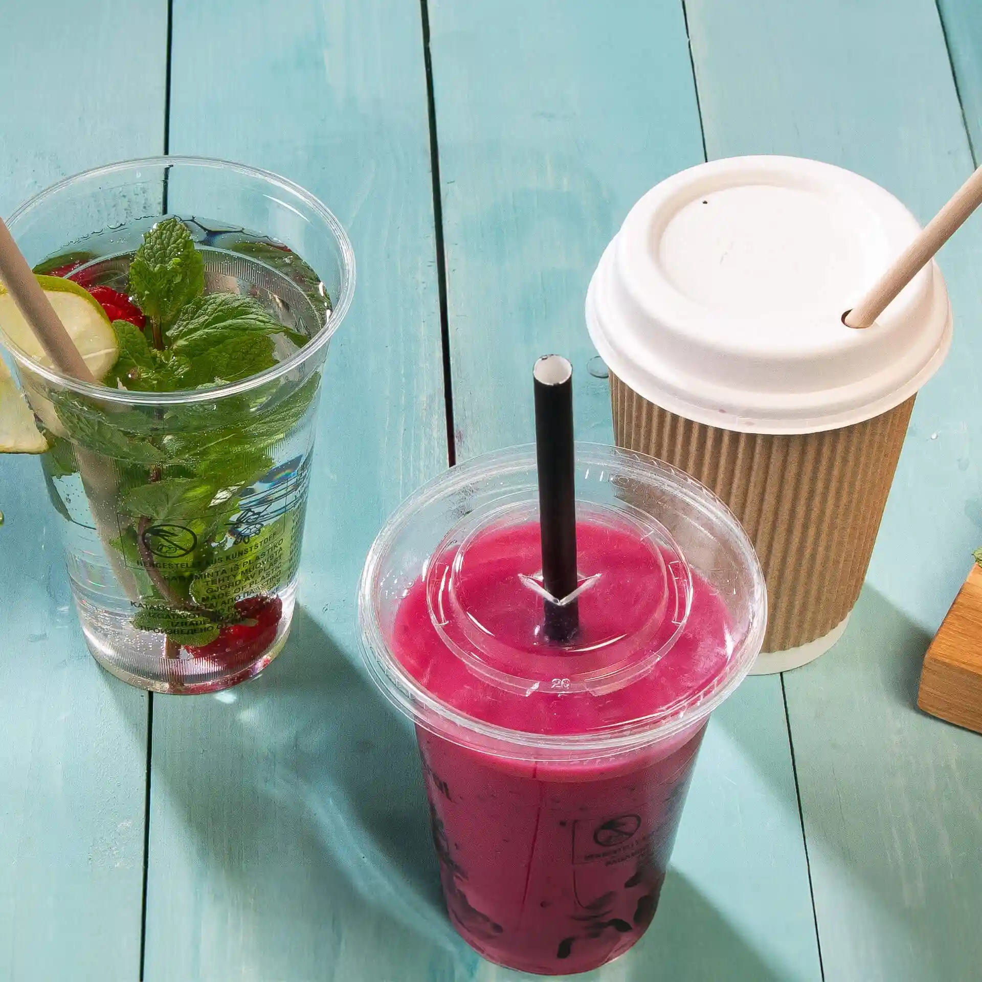 Image de trois boissons sur une surface en bois bleu : un verre transparent avec de la menthe et du citron, une boisson rose avec une paille noire et un gobelet en papier.