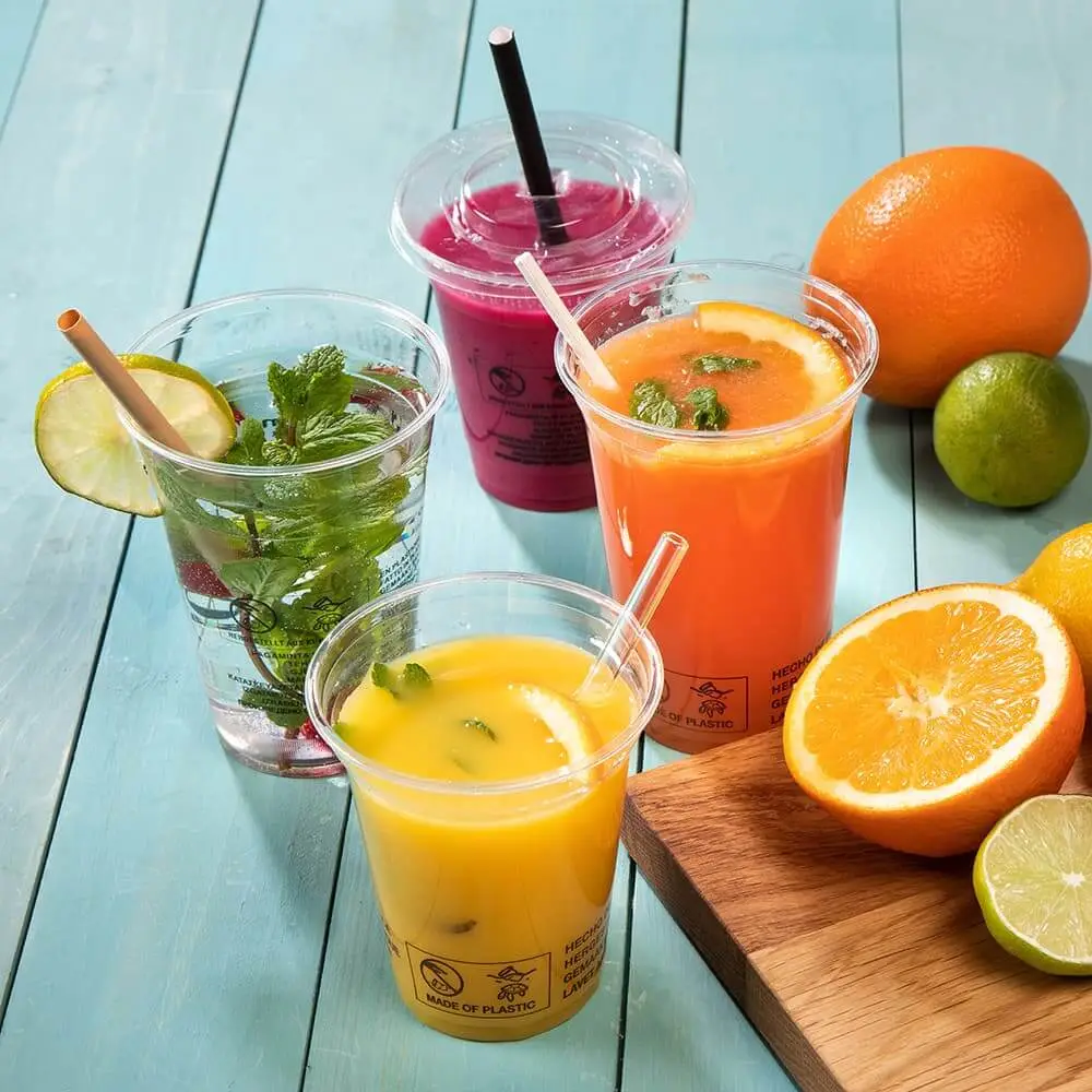 Quatre gobelets en plastique transparent remplis de boissons colorées, dont du jus d'orange et un smoothie rose, à côté de fruits frais.