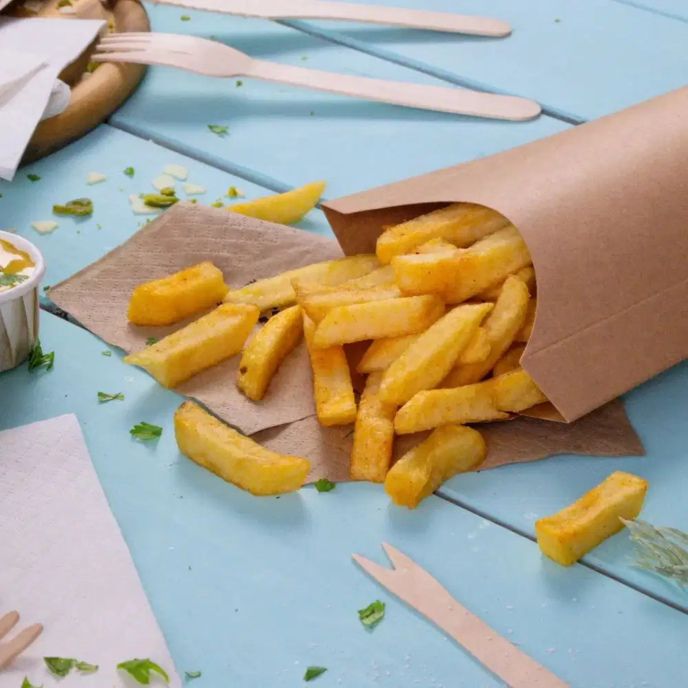 Conteneur en papier brun renversant des frites dorées sur une table bleue avec des serviettes et des couverts en bois à proximité.