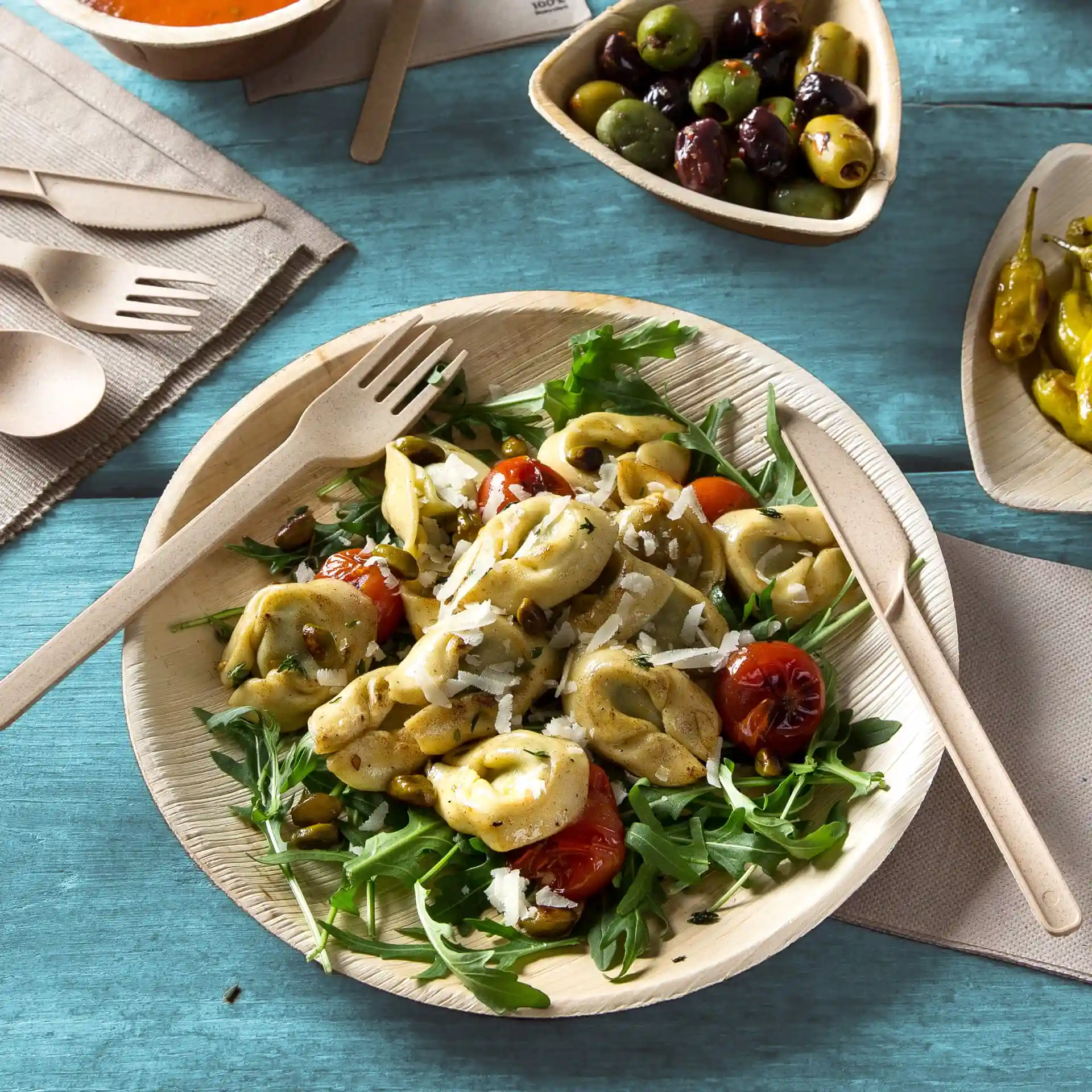 Assiette ronde en feuille de palmier avec salade de tortellini, roquette et tomates cerises, accompagnée d'ustensiles en bois sur une surface bleue.