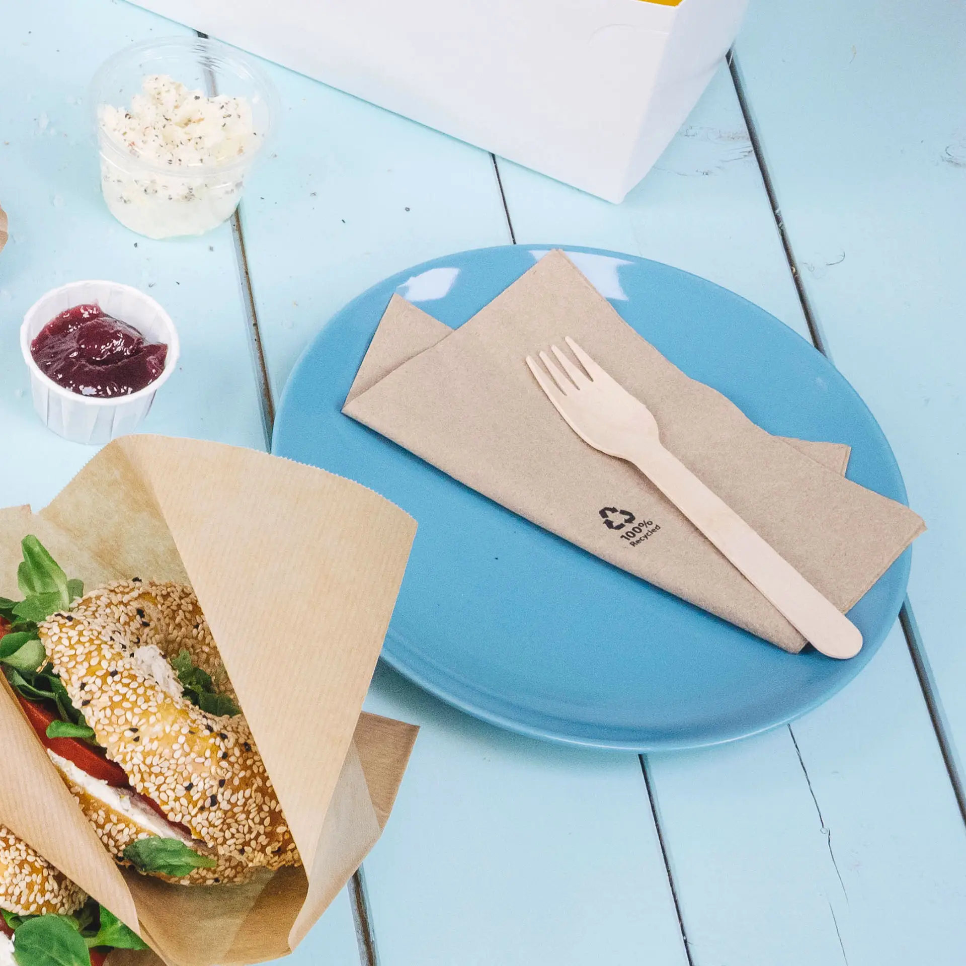 Une assiette bleue avec des serviettes en papier marron pliées et une fourchette en bois ; un bagel au sésame avec des légumes à côté, de la confiture dans une tasse.