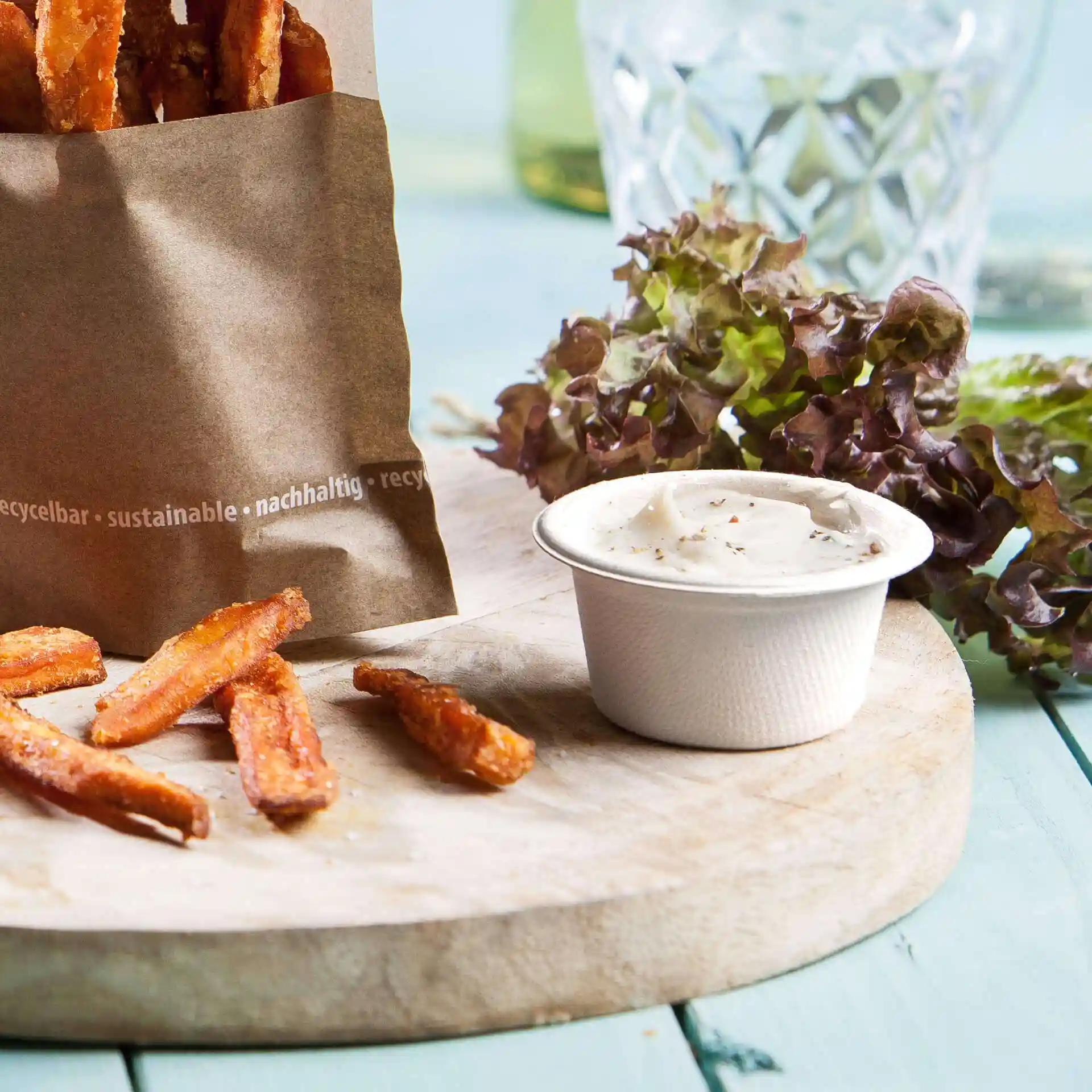 Image d'un sac en papier rempli de frites de patate douce, d'un bol de sauce blanche et de légumes verts sur une planche en bois.