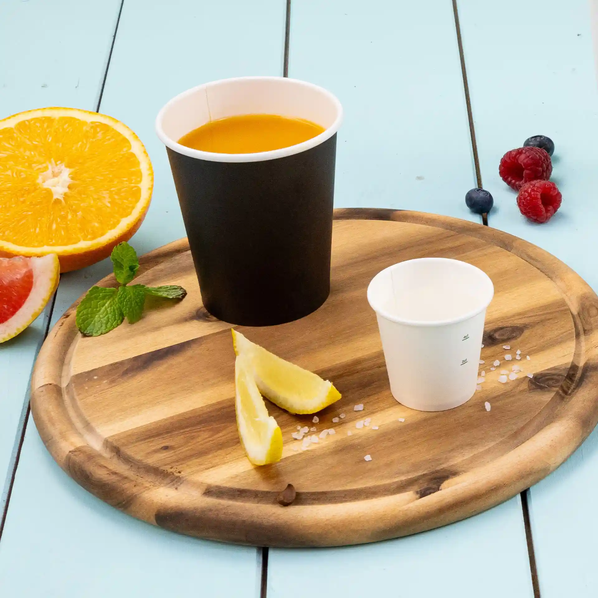 Gobelet noir avec liquide orange, petit gobelet blanc, quartiers de citron et fruits frais sur un plateau en bois.