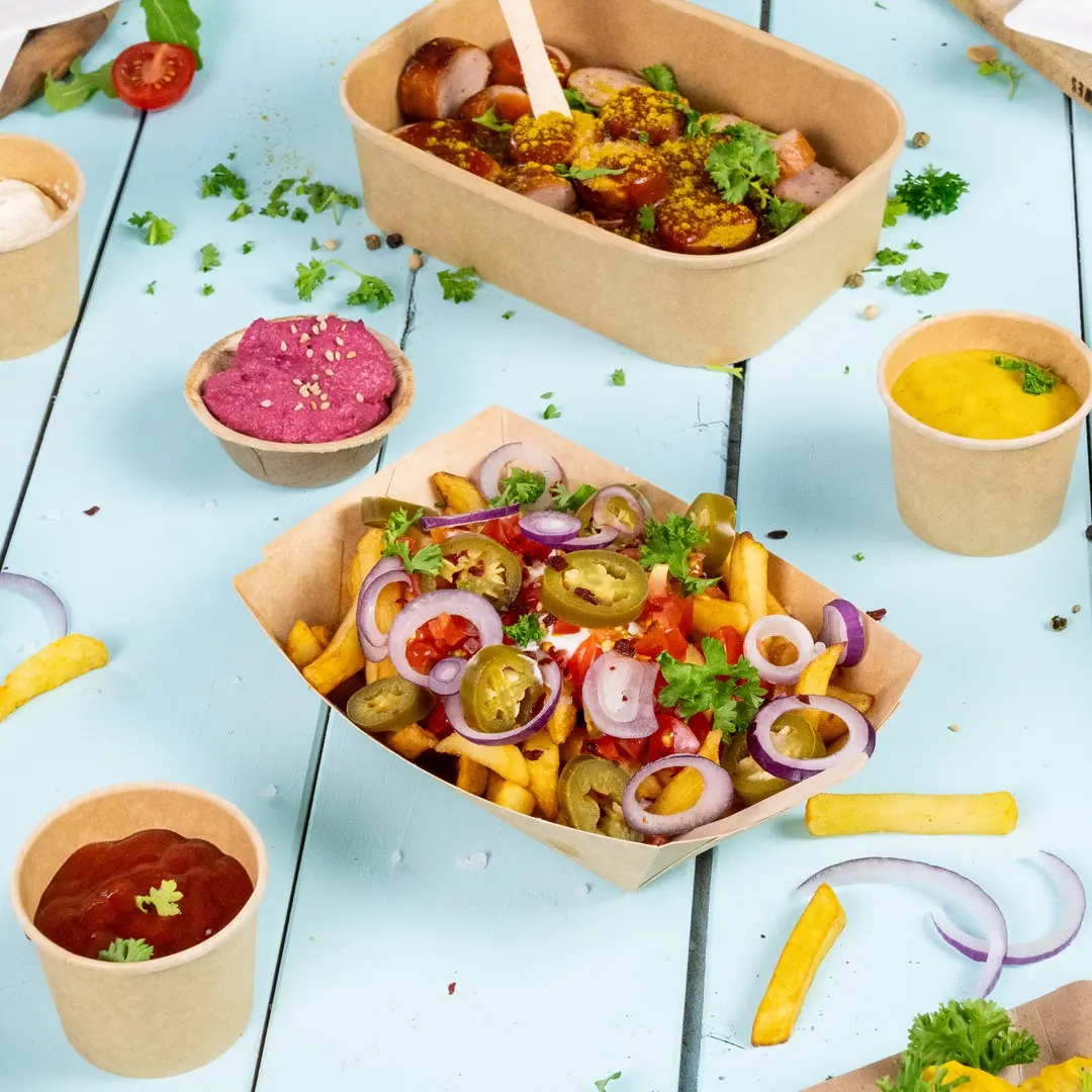 Image de divers aliments, y compris un contenant de saucisses, une boîte de frites garnies et de petits pots de sauces sur une surface en bois bleu.