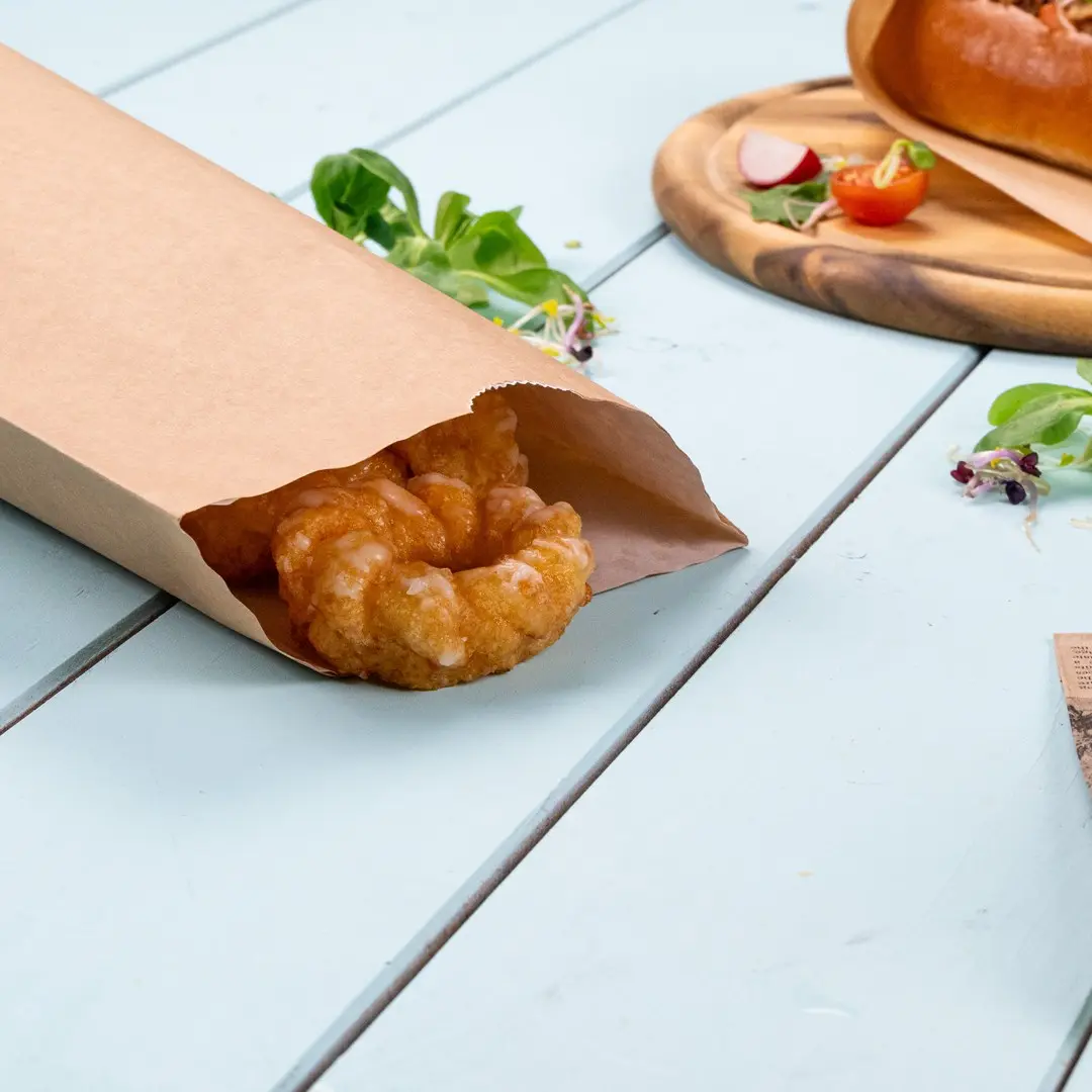 Sac en papier brun partiellement ouvert avec une pâtisserie à l'intérieur, posé sur une surface en bois bleu clair.