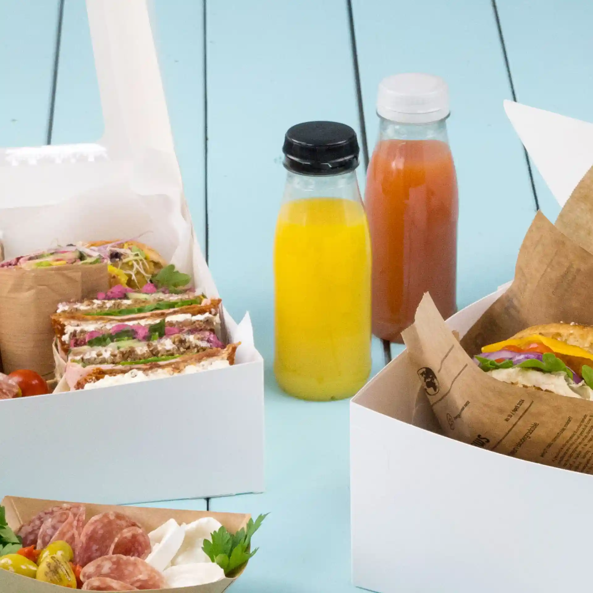 Image de contenants alimentaires avec des sandwiches, une salade et deux bouteilles transparentes avec des couvercles noirs et blancs sur une surface bleue.