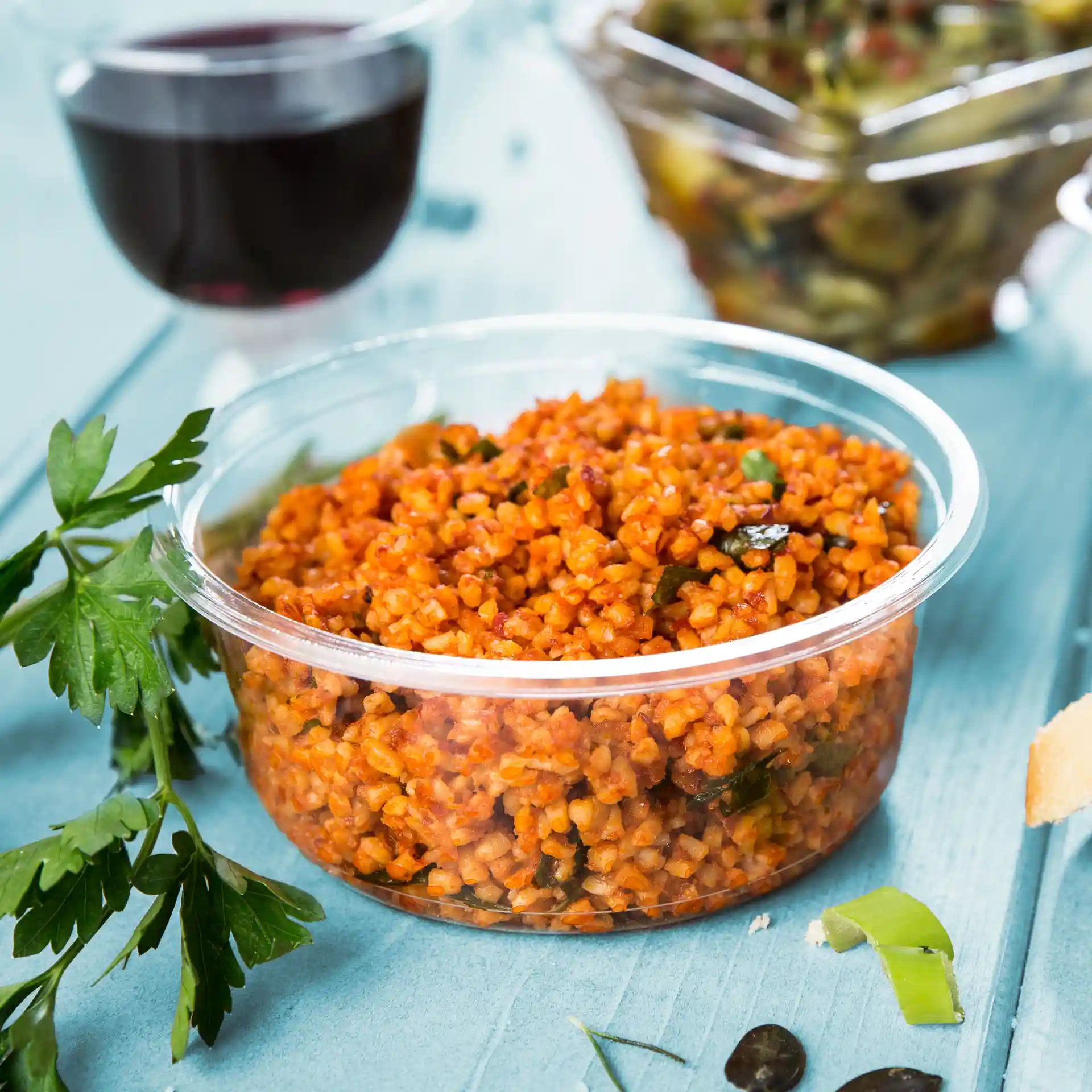 Conteneur deli transparent rond rempli de bulgur orange, garni de persil, sur une surface bleue.