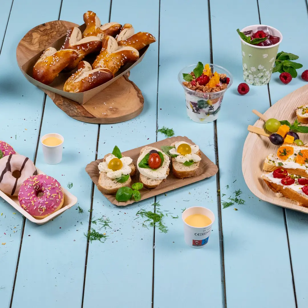 Une variété de finger foods comprenant des bretzels, des sandwiches garnis, des beignets et des coupes de fruits sur une table en bois bleue.