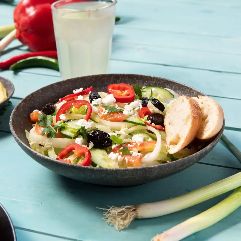 Bol de salade avec concombre, tomates, olives, fromage feta et tranches de poivron rouge, accompagné de deux morceaux de pain.