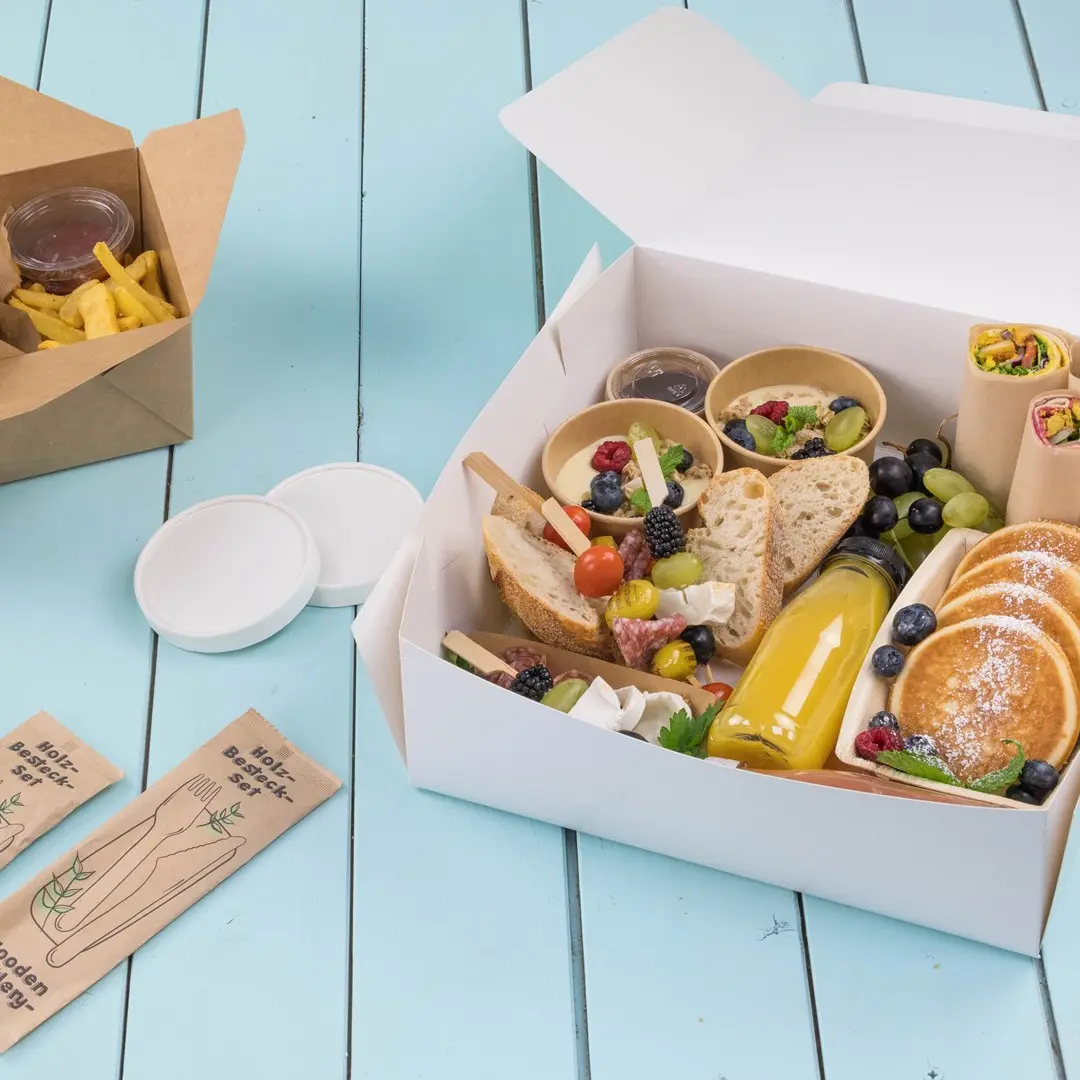 Boîte à emporter blanche remplie de divers aliments, y compris du pain, des fruits, des crêpes et une boisson, sur une surface en bois bleu.