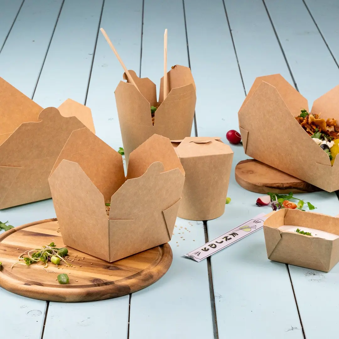 Contenants alimentaires en carton brun de différentes formes sur une planche en bois avec des ingrédients frais éparpillés.