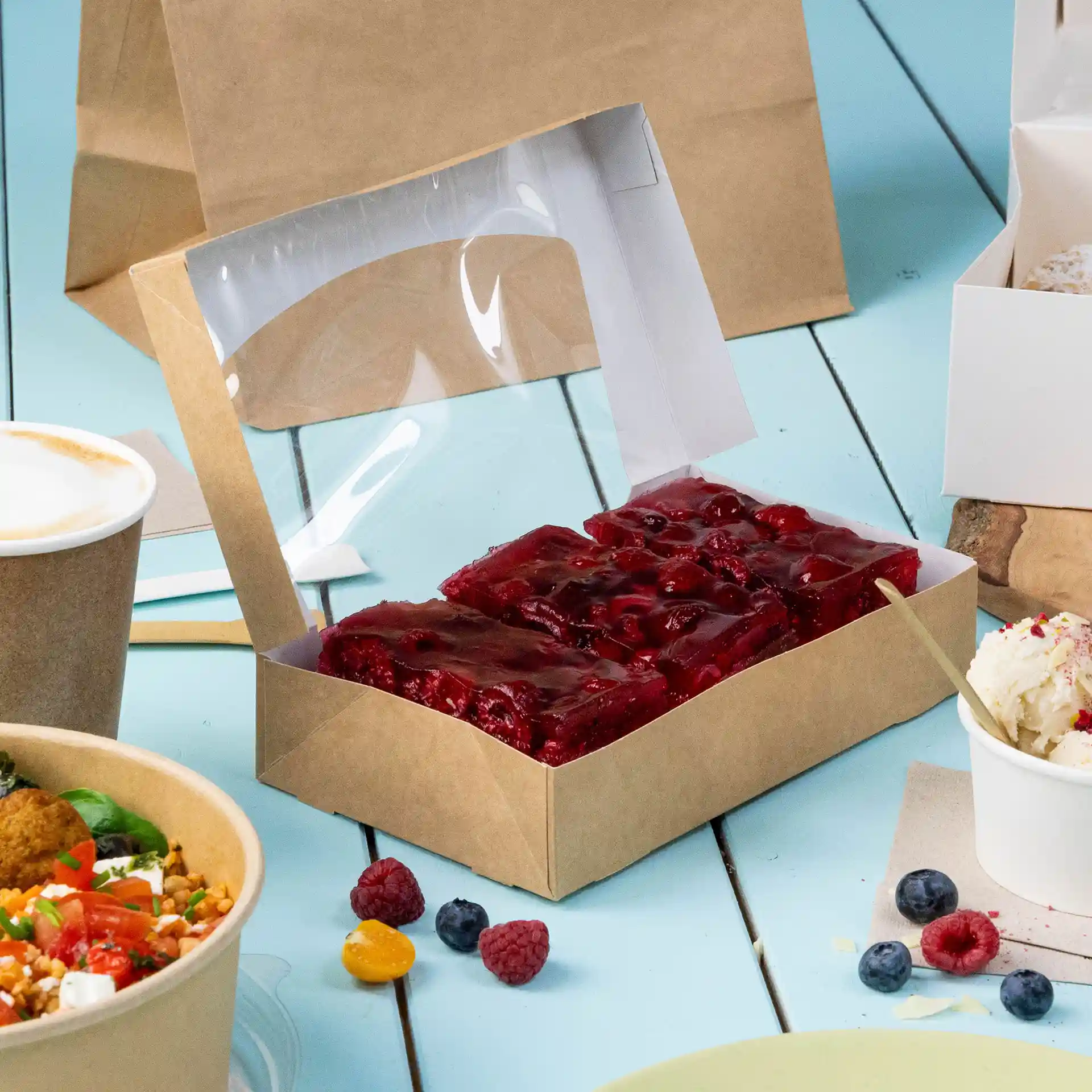 Boîte en carton avec couvercle transparent, contenant un dessert en gelée rouge, sur une surface bleue avec d'autres aliments.