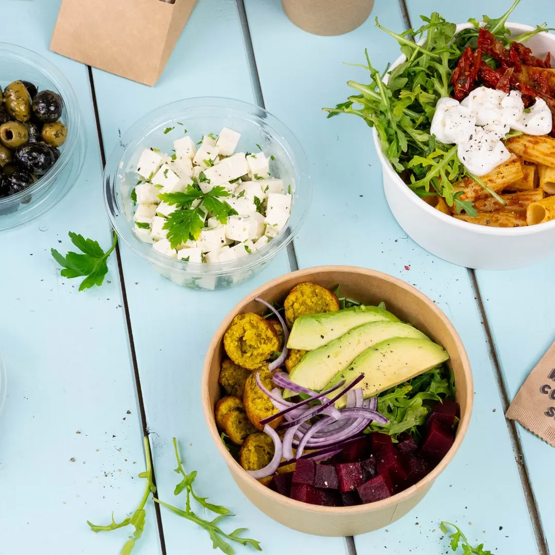 Bol avec falafel, avocat, betteraves, oignon rouge et légumes verts, à côté de contenants de feta et d'olives sur une surface bleue.