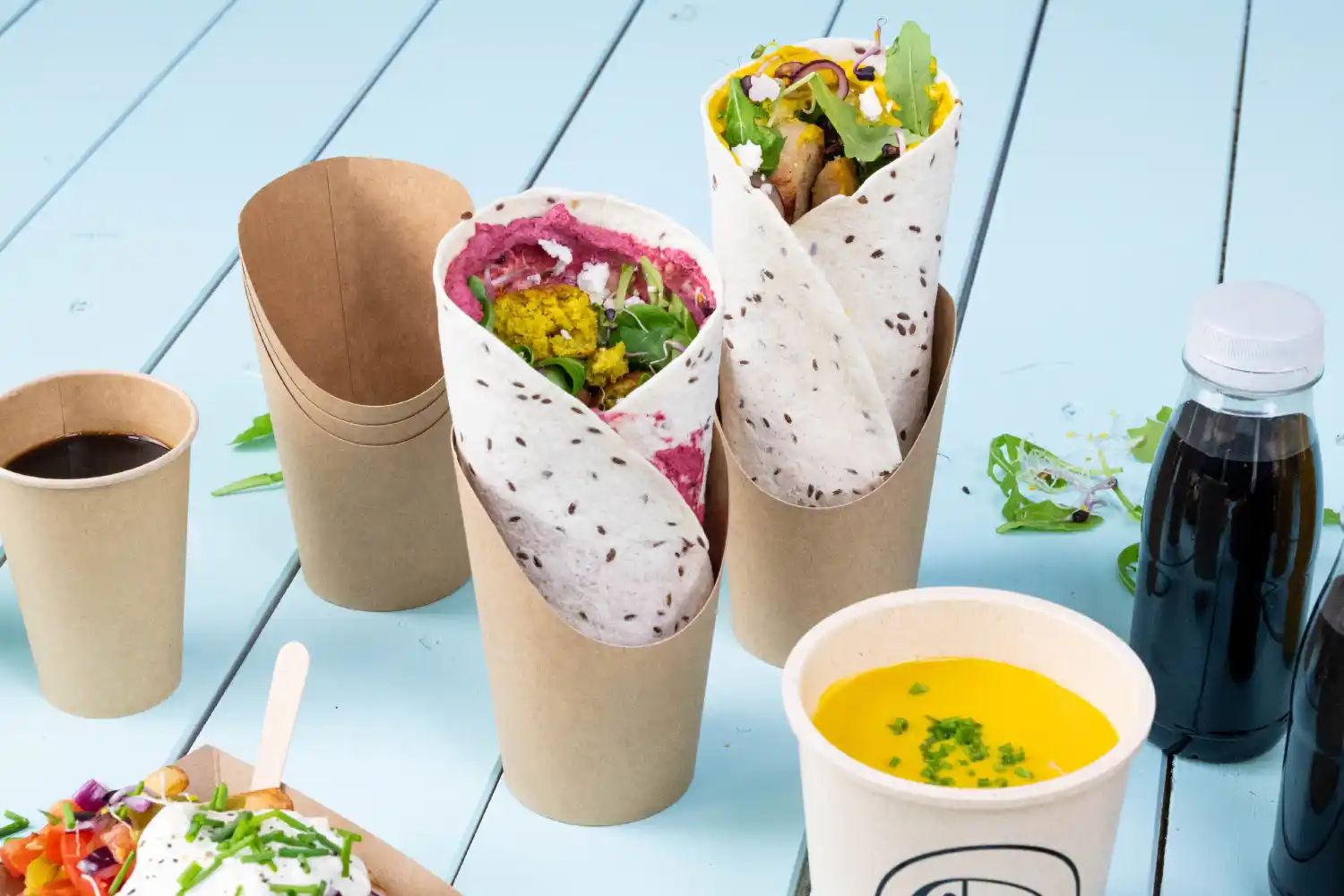 Deux aliments enveloppés dans des gobelets, l'un avec une garniture rose et des légumes, l'autre jaune, à côté d'une tasse de café et de soupe.