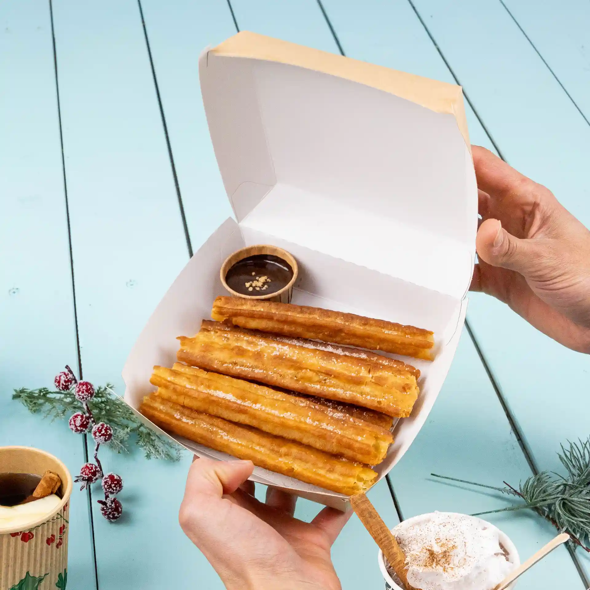 Boîte à emporter contenant plusieurs churros avec un petit pot de sauce au chocolat, sur une surface en bois bleu clair.