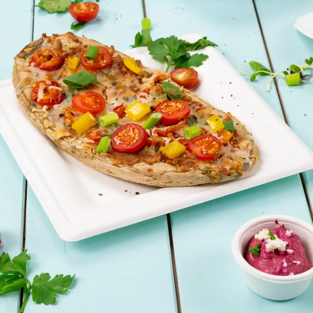 Pain plat cuit garni de tomates cerises, oignons verts et poivrons, servi sur une assiette blanche avec un accompagnement de sauce.