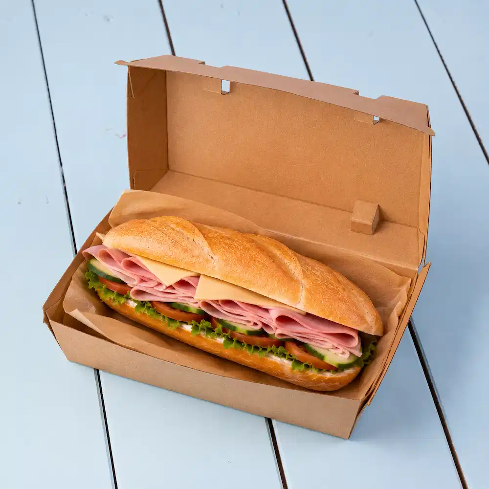 Sandwich dans une boîte à emporter marron, avec des couches de jambon, de fromage, de laitue et de concombre tranché.