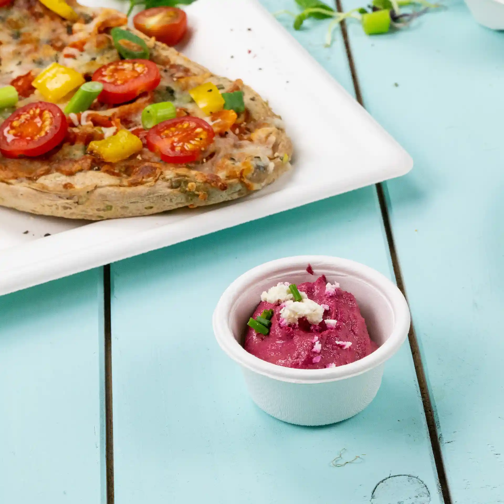 Bol en bagasse blanc avec une sauce rose et une garniture verte, placé à côté d'une pizza sur une assiette blanche.