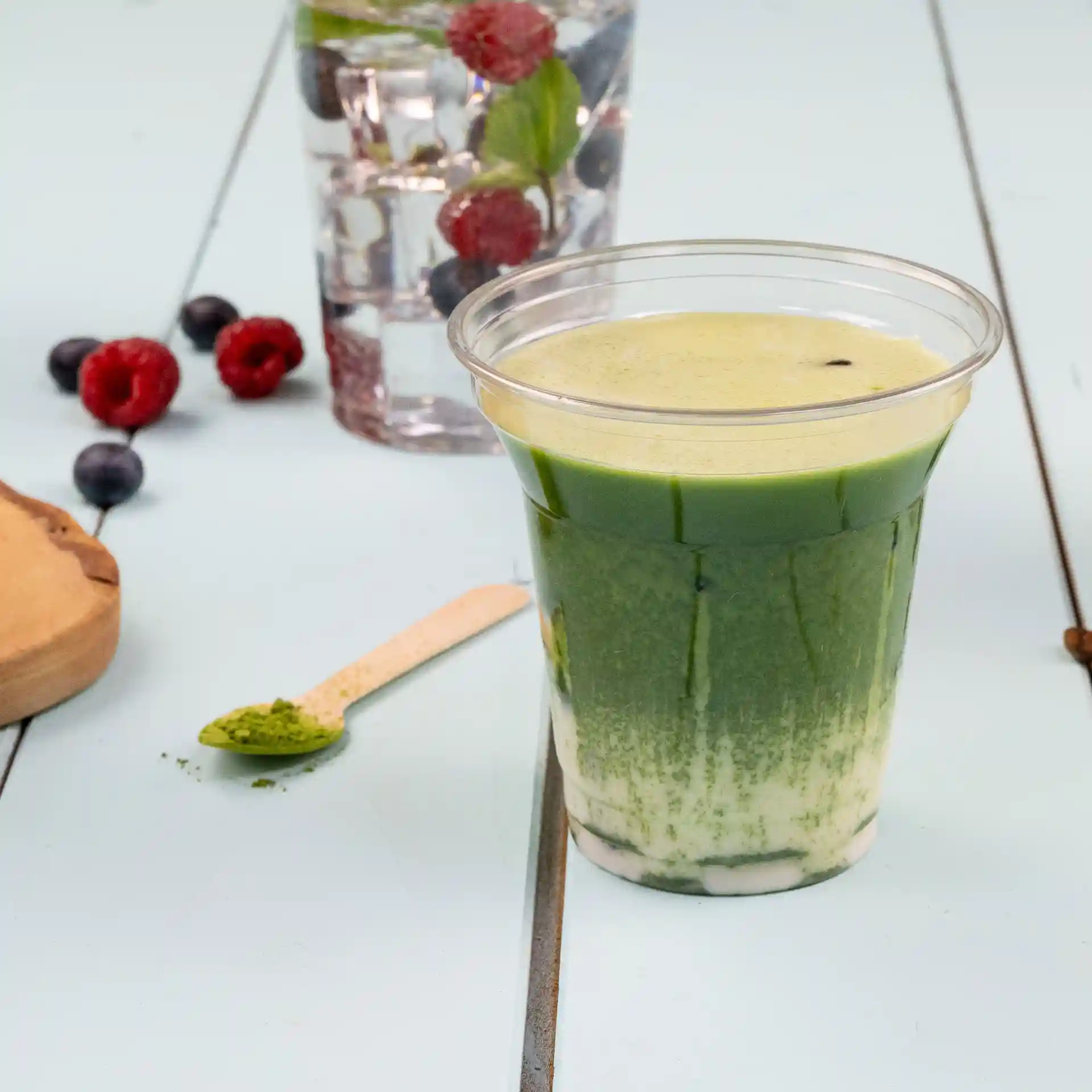 Gobelet rPET transparent rempli de boisson verte, avec une cuillère en bois et des baies en arrière-plan.