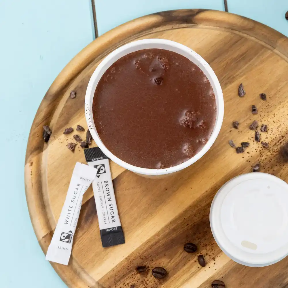 Chocolat chaud dans une tasse blanche avec des sachets de sucre, sur un plateau en bois avec des grains de café et du cacao renversé.