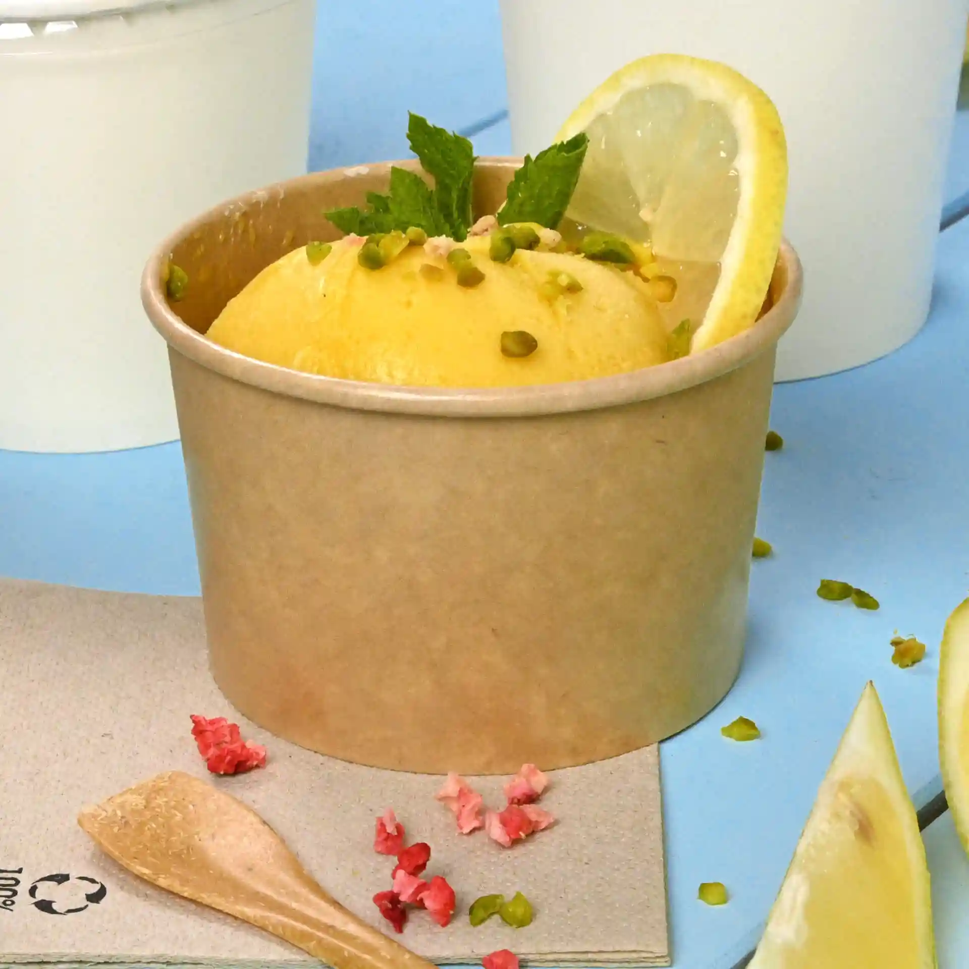 Gobelet en carton marron rempli de glace jaune, surmonté d'une tranche de citron et de menthe, sur une surface bleue avec une cuillère en bois.