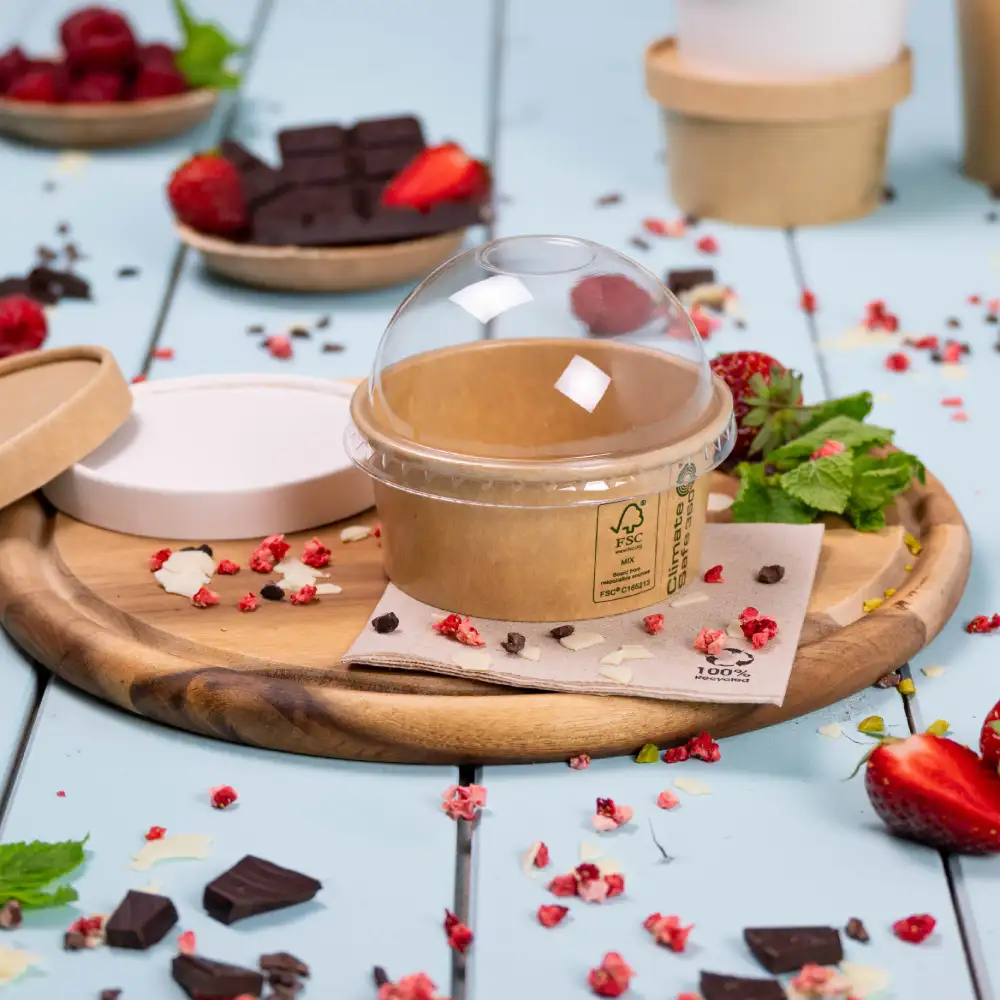 Contenant à dessert à emporter avec un couvercle transparent sur un plateau en bois, entouré de fraises fraîches, de framboises et de morceaux de chocolat.