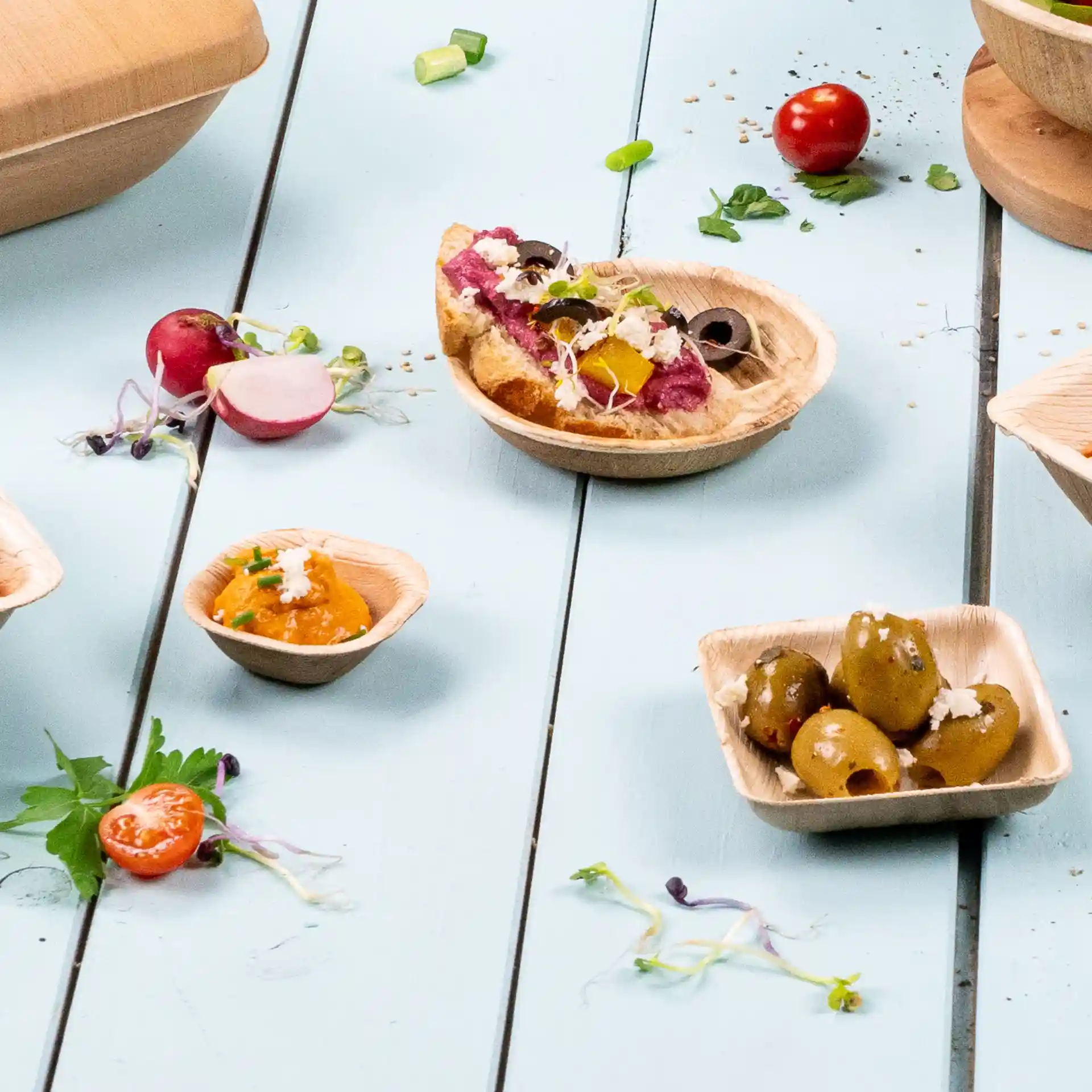 Image de bols en feuille de palmier avec divers aliments, y compris des olives, un dip et un petit sandwich, sur une surface en bois bleu.