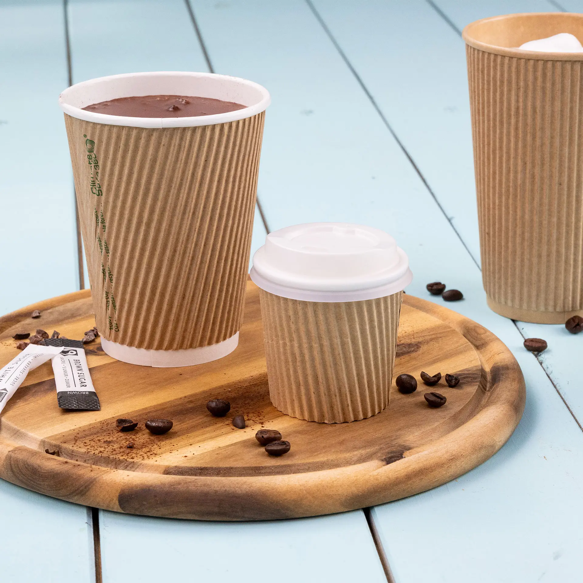 Une tasse à café marron nervurée avec du chocolat chaud, une tasse plus petite avec un couvercle blanc et une plus grande tasse à côté sur un plateau en bois.