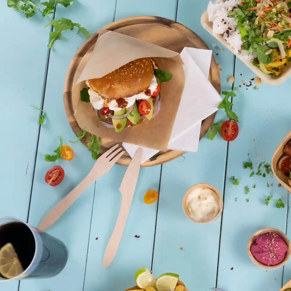 Une assiette en bois contient un burger aux graines de sésame avec des légumes, accompagné de couverts et divers aliments sur fond bleu.
