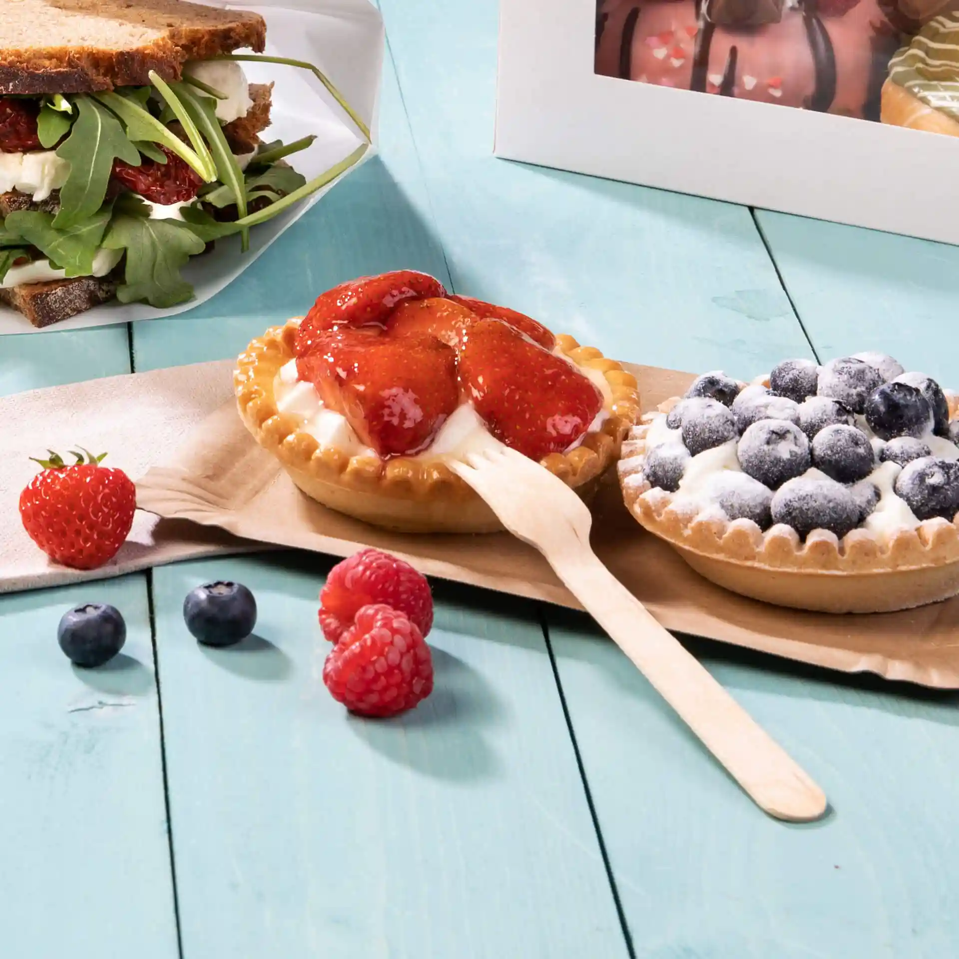 Image de deux tartes dessert avec des garnitures de fraises et de myrtilles, accompagnées de baies fraîches et d'une fourchette en bois sur une surface bleu clair.