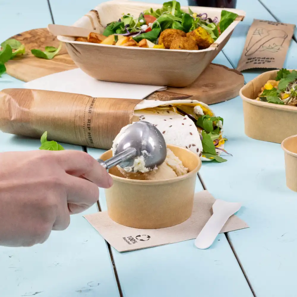 Une main utilisant une cuillère à glace en métal pour servir d'un gobelet en papier brun ; d'autres aliments et contenants sont en arrière-plan sur une table bleue.