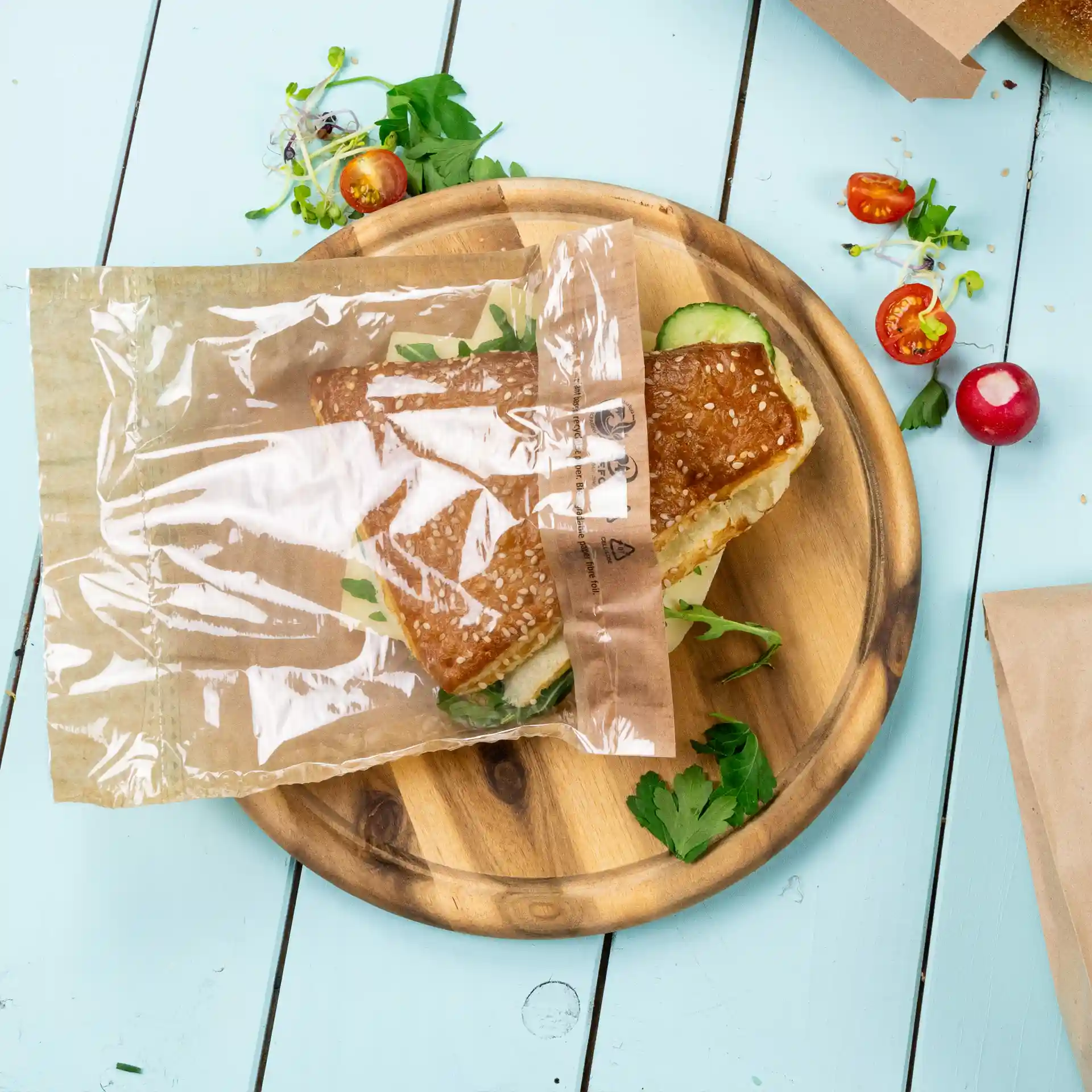 Sac à collation transparent avec un sandwich à l'intérieur, placé sur une assiette en bois avec des herbes et des légumes frais.