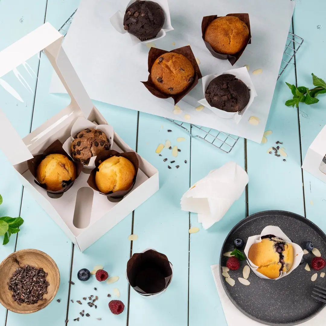 Une boîte à cupcakes blanche contenant trois muffins, entourée de muffins supplémentaires, de baies et de morceaux de chocolat sur une surface bleue.