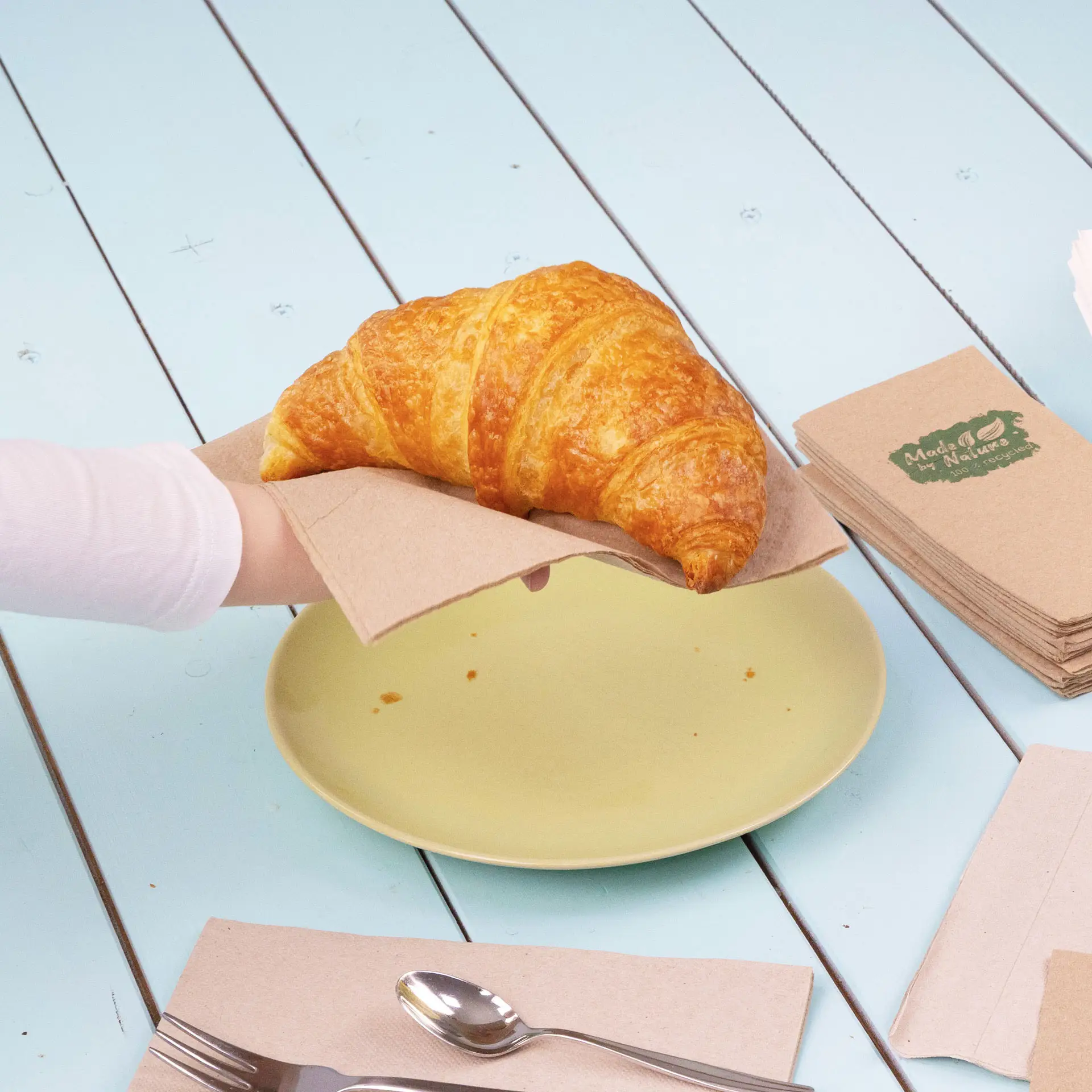 Une main tenant un croissant sur une serviette en papier au-dessus d'une assiette jaune, avec des serviettes et des couverts à proximité.