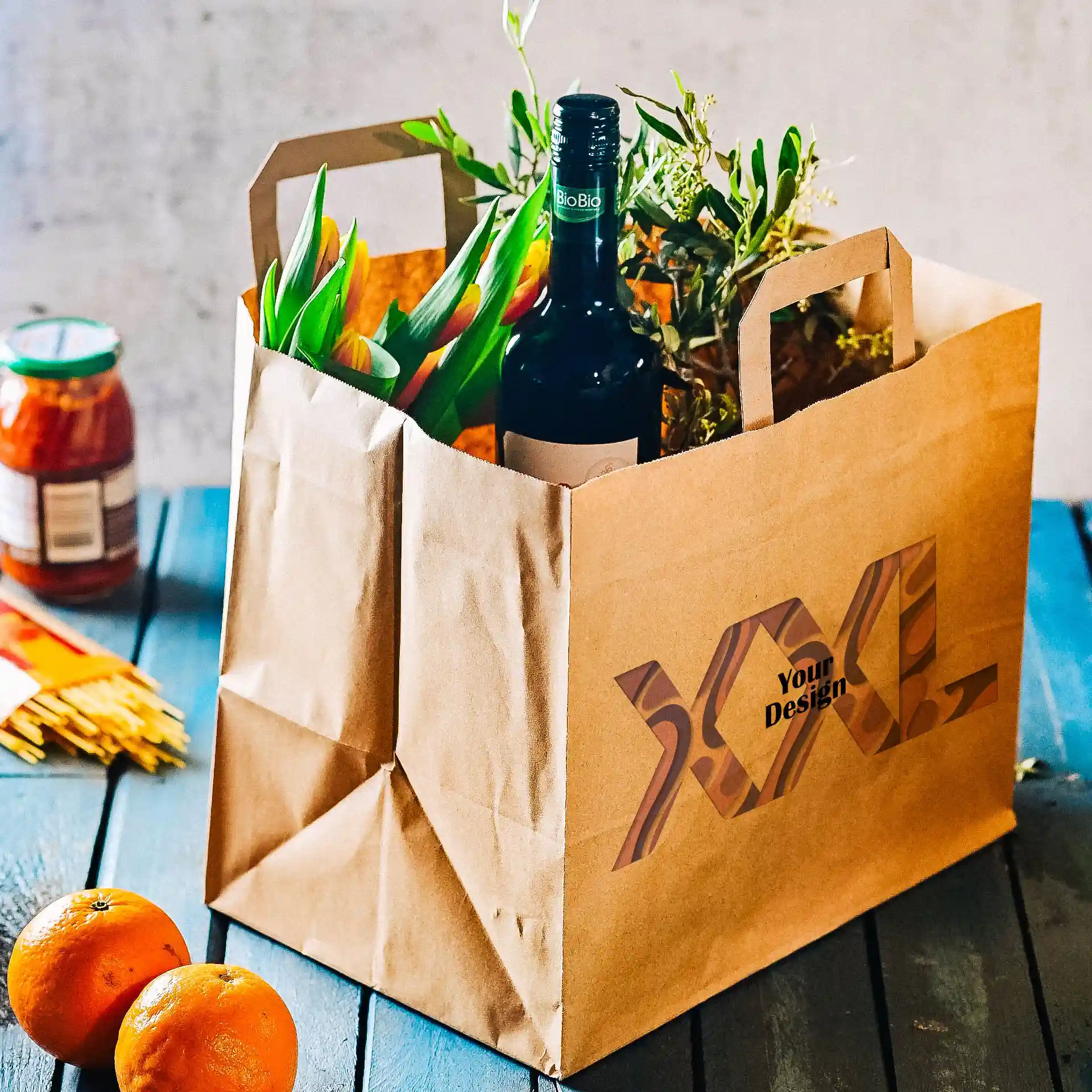 Sac en papier brun avec poignées, rempli de fleurs, d'une bouteille et de verdure, sur une surface en bois bleu.