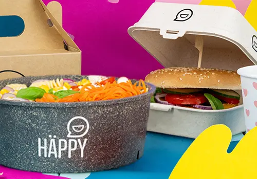 Empilement de bols colorés étiquetés "HAPPY BOWL" dans divers designs, avec un contenant alimentaire séparé en arrière-plan.