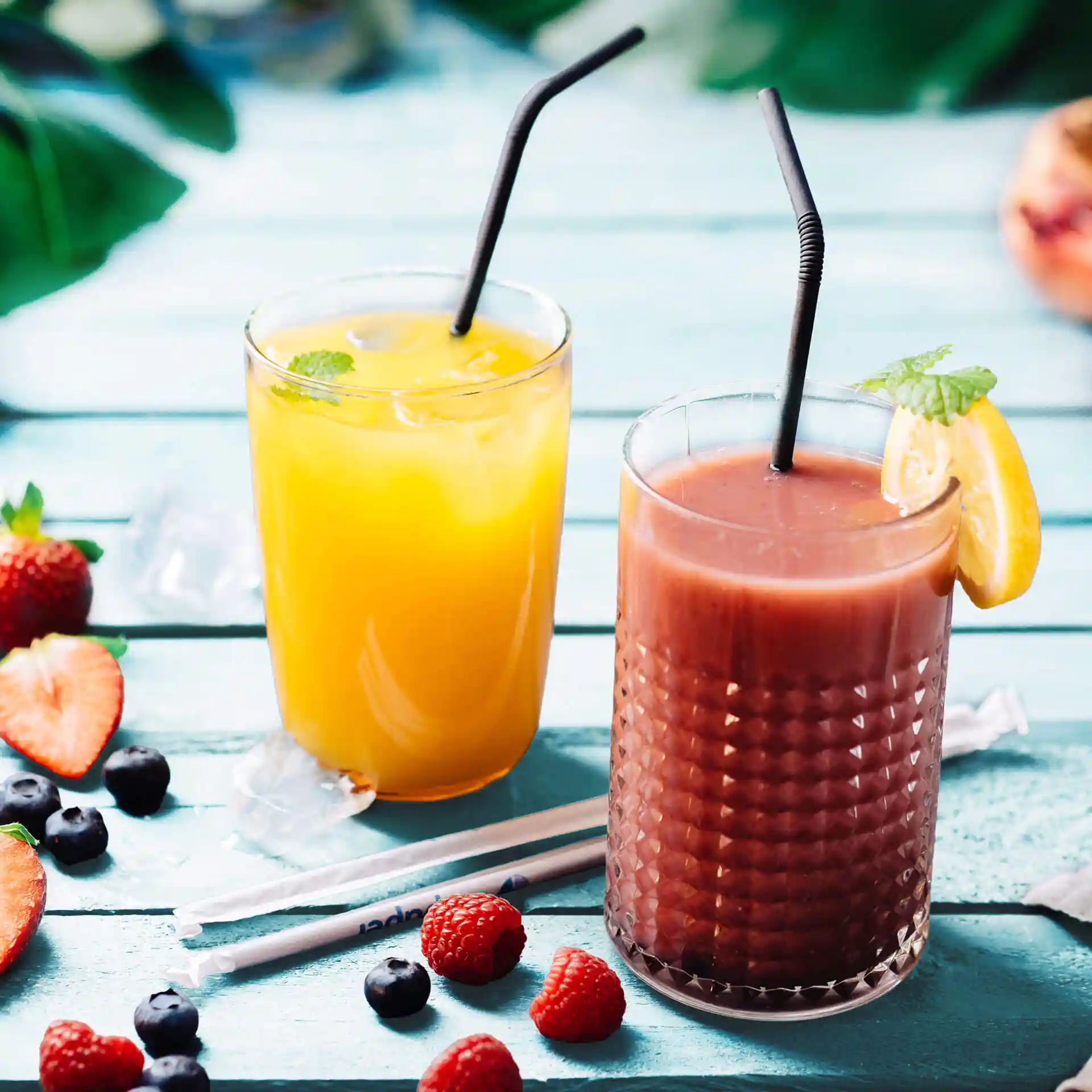 Image de deux boissons dans des verres avec des pailles noires, entourées de fraises, myrtilles et framboises sur une surface bleue.