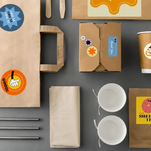 Une collection d'emballages alimentaires jetables, comprenant un sac en papier, des boîtes, des gobelets et des ustensiles avec des étiquettes et autocollants colorés.