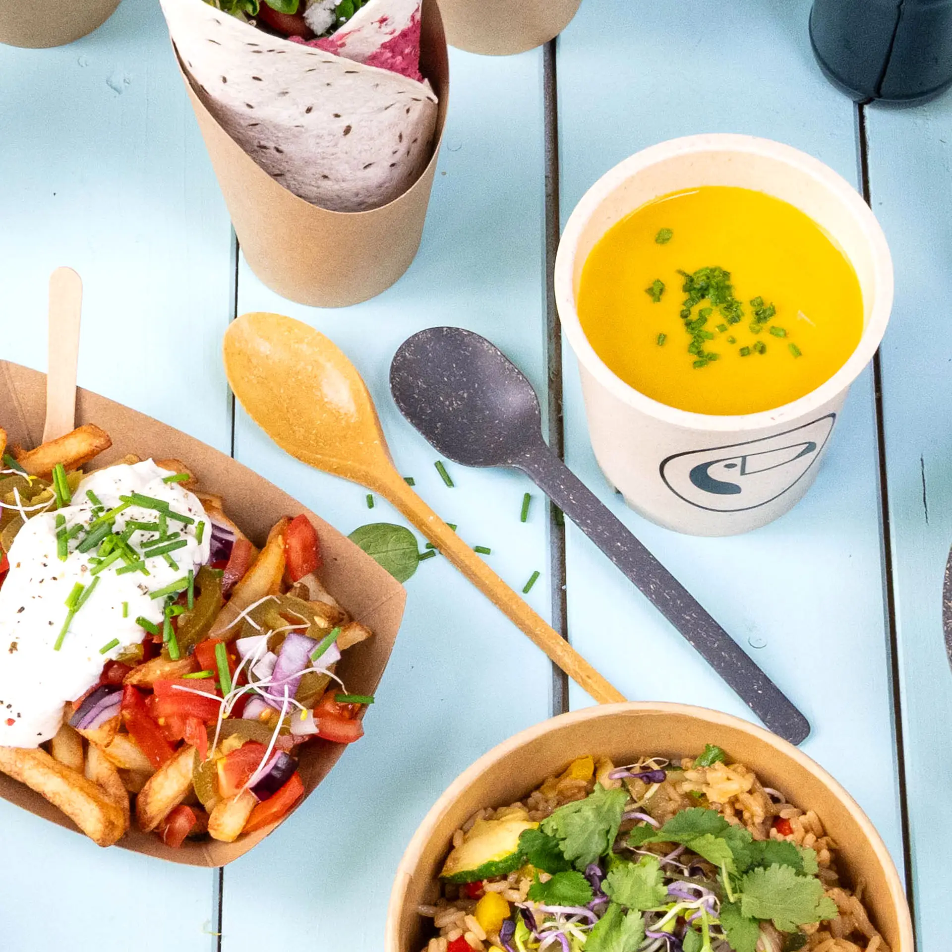 Une cuillère réutilisable couleur caramel en bio- composé ; inclut divers aliments et une tasse à soupe jaune avec de la ciboulette.