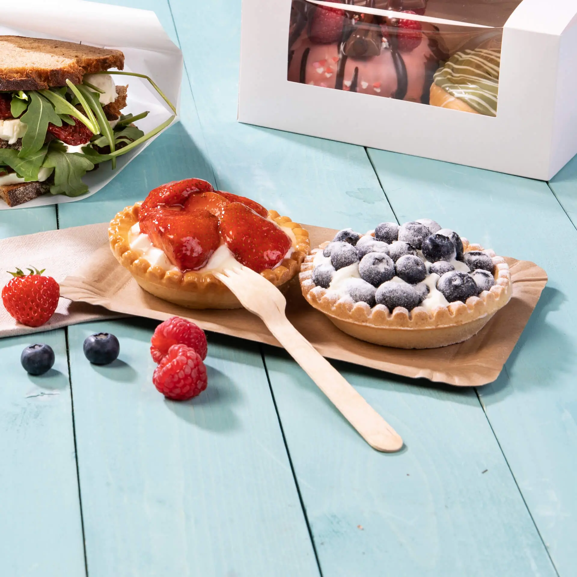 Deux tartes dessert avec des fraises et des myrtilles sur une assiette en carton marron, accompagnées de baies fraîches et d'un sandwich.