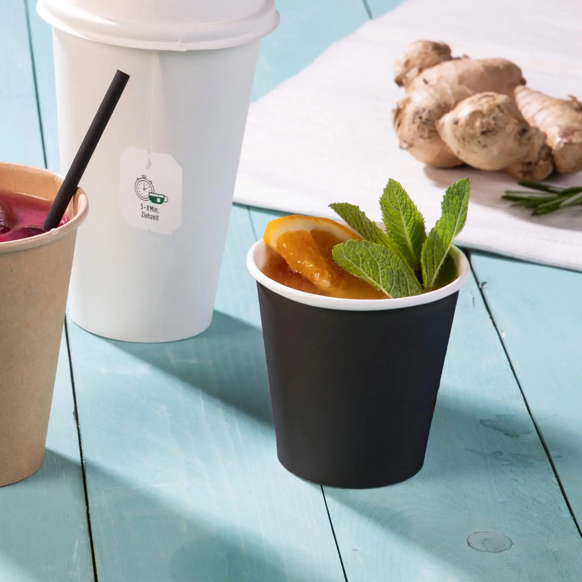 Gobelet en papier noir à paroi simple avec revêtement PLA, rempli de tranches d'orange et de feuilles de menthe, à côté d'autres gobelets et de gingembre.