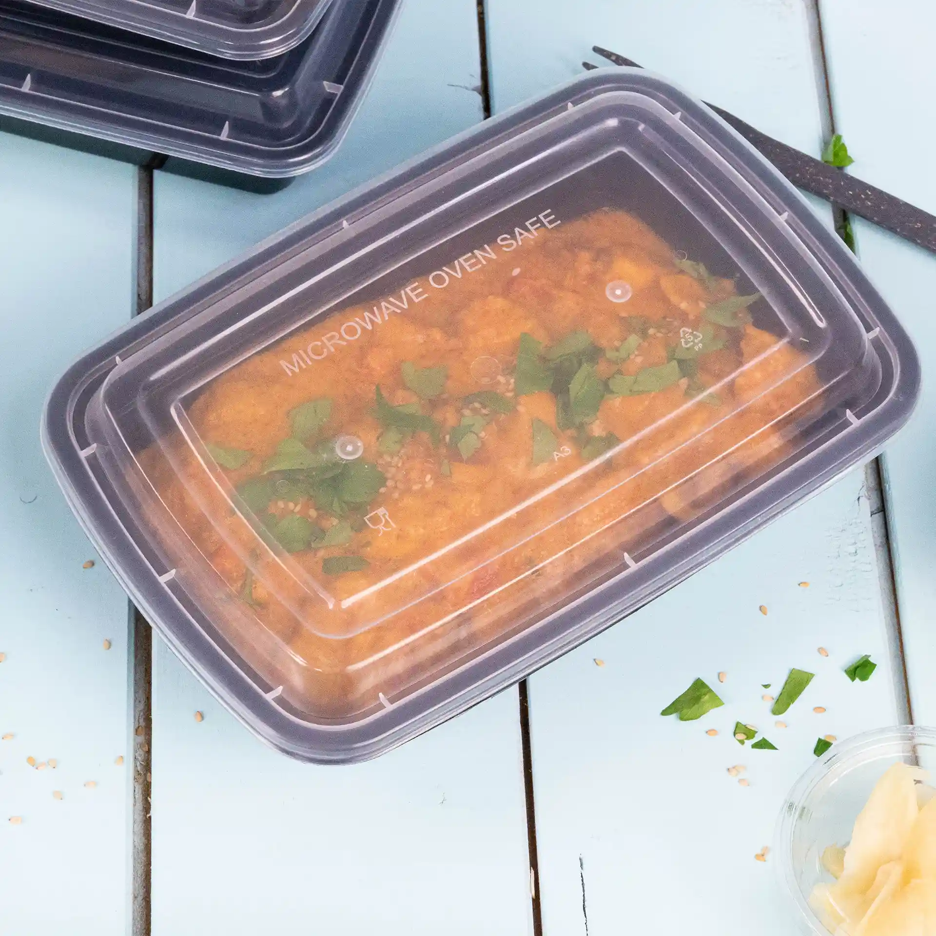 Conteneur rectangulaire transparent avec un couvercle réutilisable, rempli de nourriture et garni d'herbes vertes, sur une surface bleue.