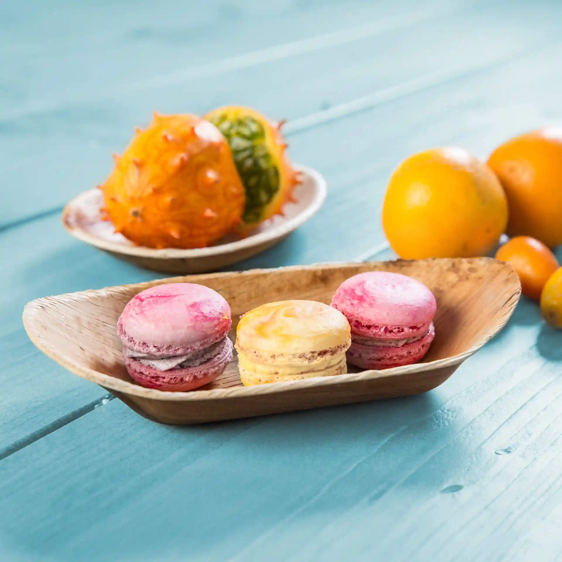 Plat en forme de bateau en bambou avec des macarons roses et jaunes, entouré de divers fruits sur une surface en bois bleu.