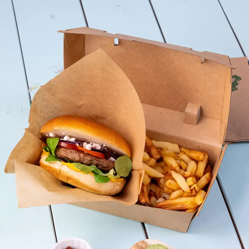 Burger dans un emballage en papier avec des légumes frais à côté d'une boîte de frites sur une surface bleue.