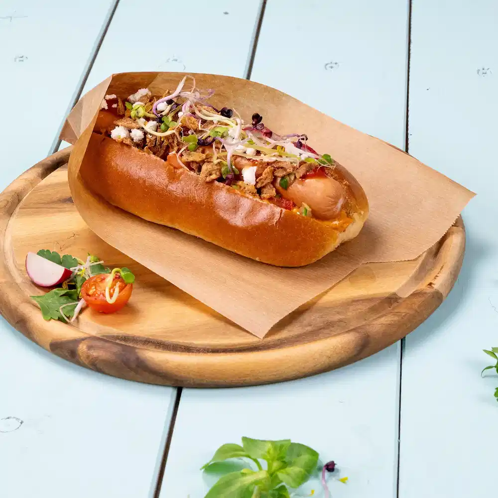 Hot dog dans un pain moelleux garni d'oignons croustillants, de pousses et décoré de tomates cerises et de radis sur une assiette en bois.