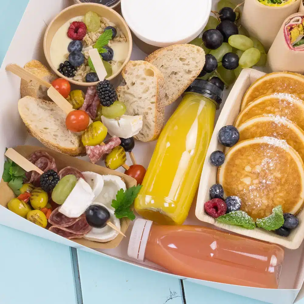 Une variété d'aliments, y compris des crêpes, des fruits, du pain et des boissons, présentée dans un récipient sur une surface bleu clair.