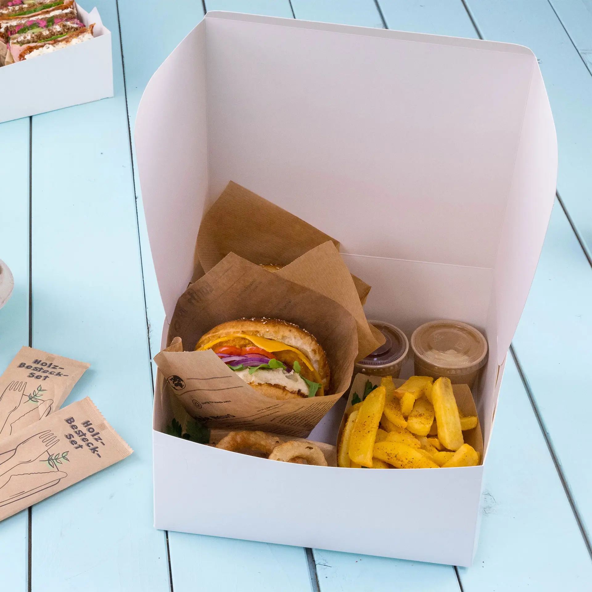 Boîte à gâteau en carton blanc contenant un sandwich, des frites et deux sauces, placée sur une surface bleu clair.