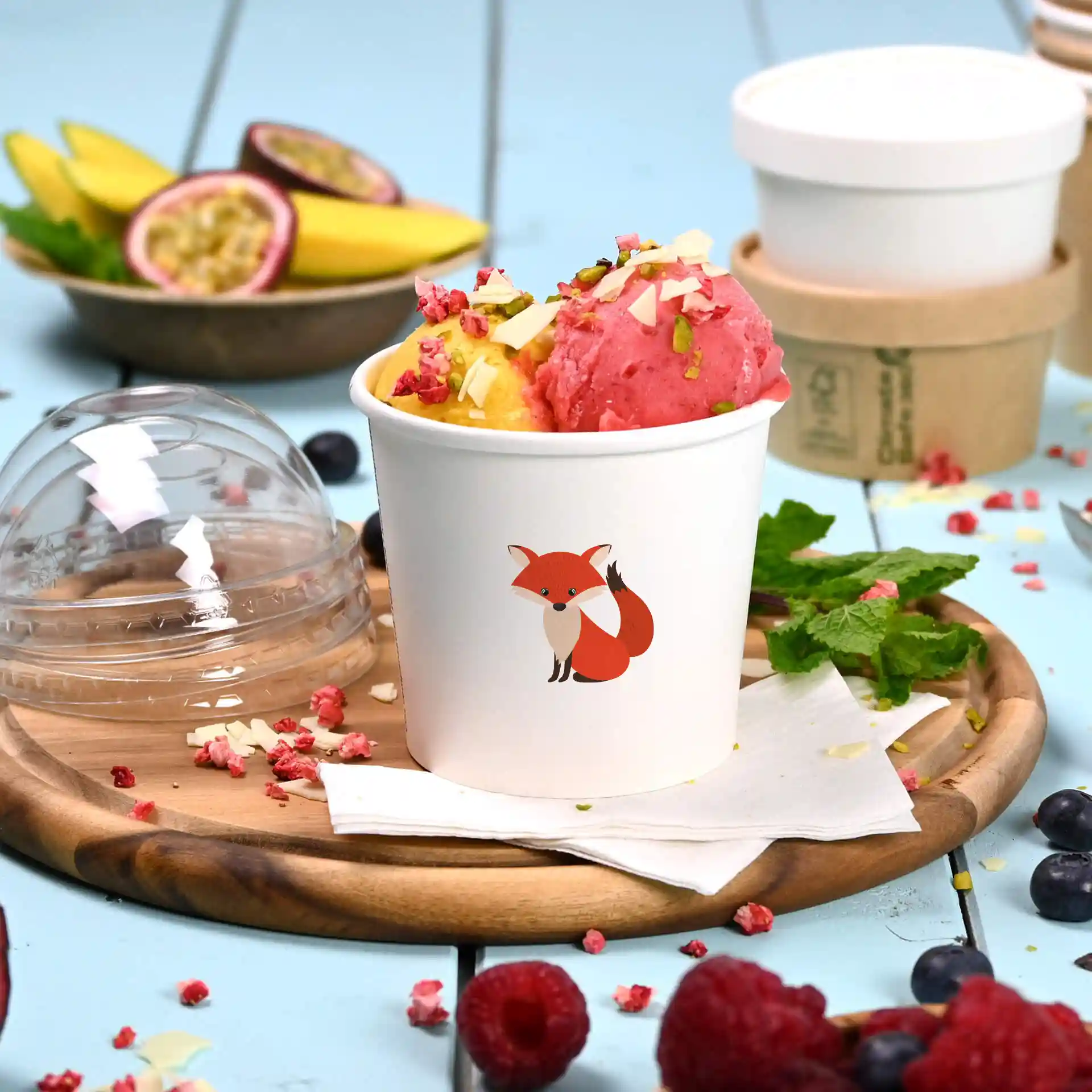 Gobelet en carton blanc pour glace avec illustration de renard, rempli de glace rose et jaune, sur une assiette en bois.