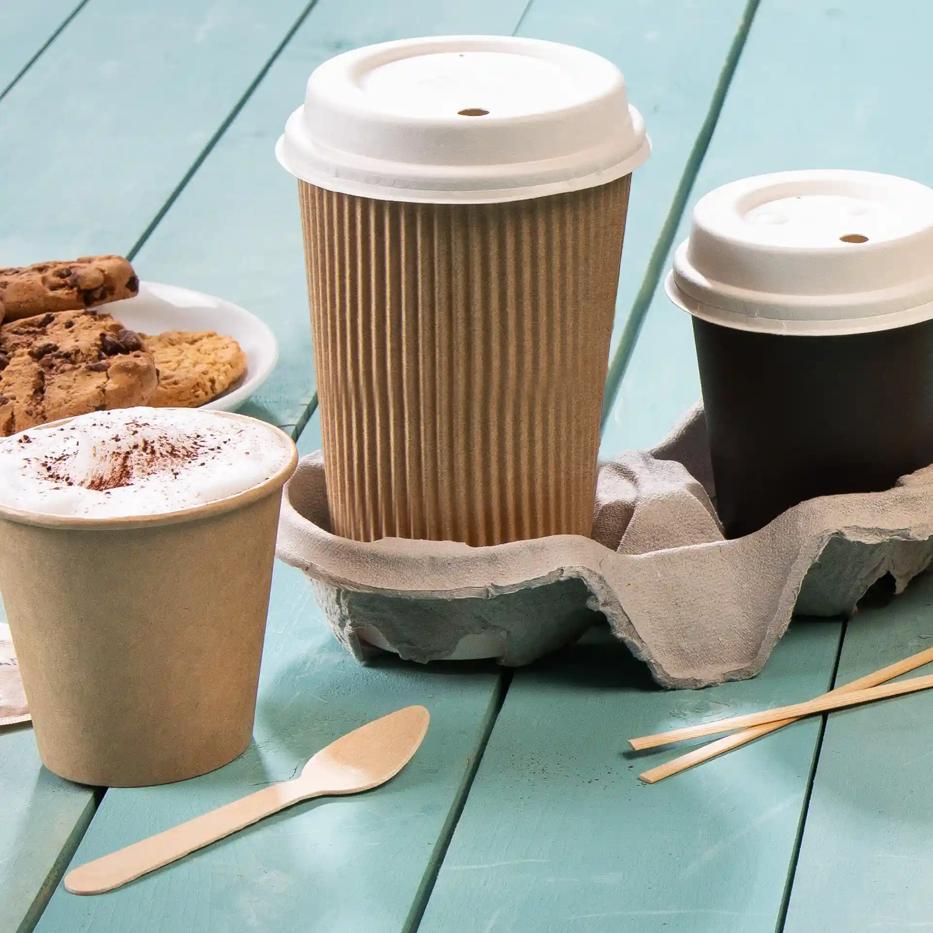Image de trois gobelets dans un support en carton, avec une assiette de biscuits et une cuillère en bois sur une table bleue.