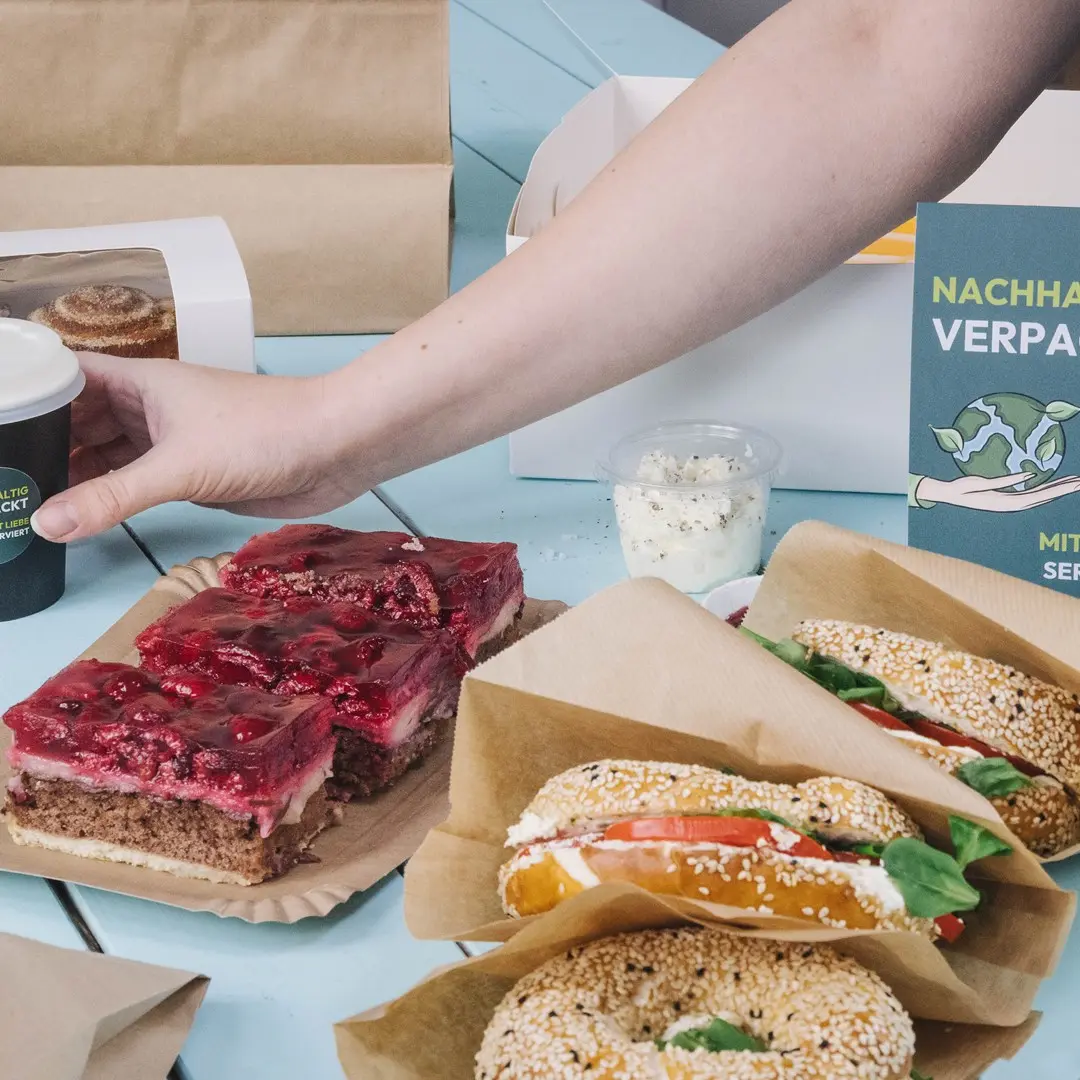 Une main tendue vers une tasse à café à côté d'une assiette de dessert en couches, deux bagels au sésame avec garnitures et un contenant de crème.